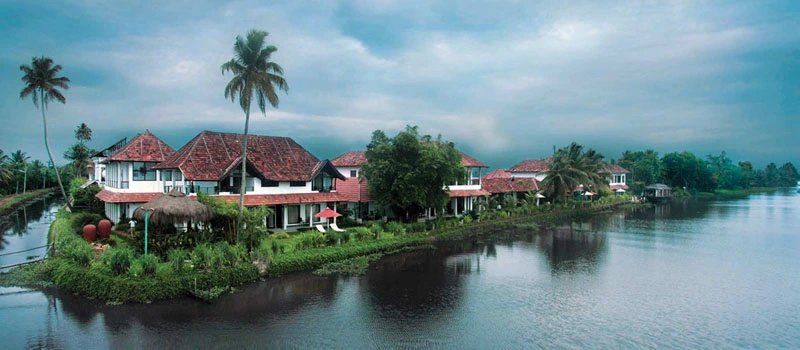 Riverside houses with red roofs among palm trees under a cloudy sky.